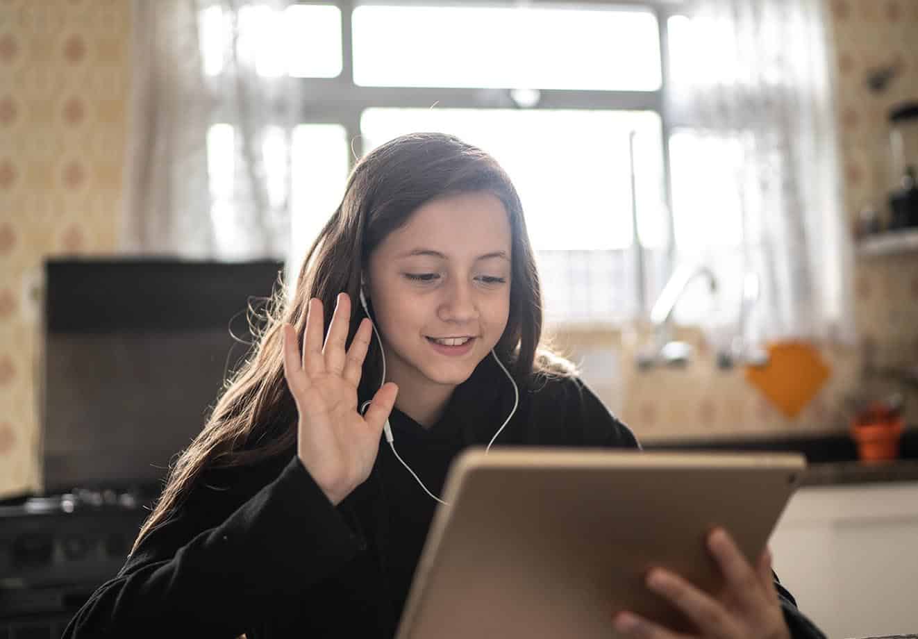 a young student sits at home in her room and waves at her online class using her ipad
