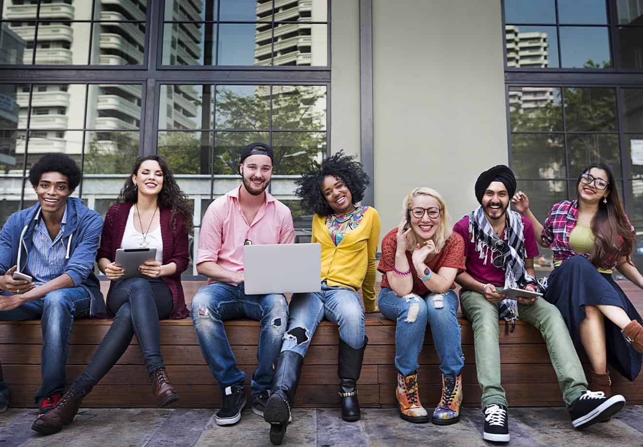 students sit on a bench outside their college building and smile at the camera