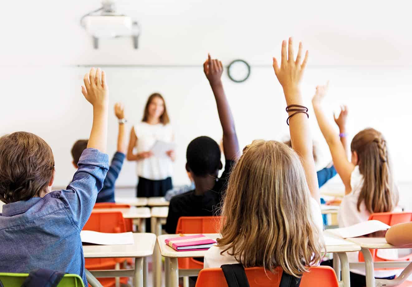 Classroom full of school children with their hands raised for the teacher at the front