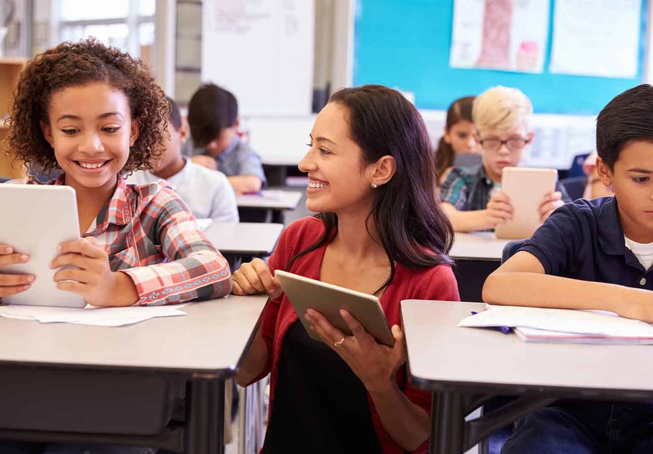 a teacher bends down to talk to one of the students in her classroom who is doing independent work in a blended learning environment