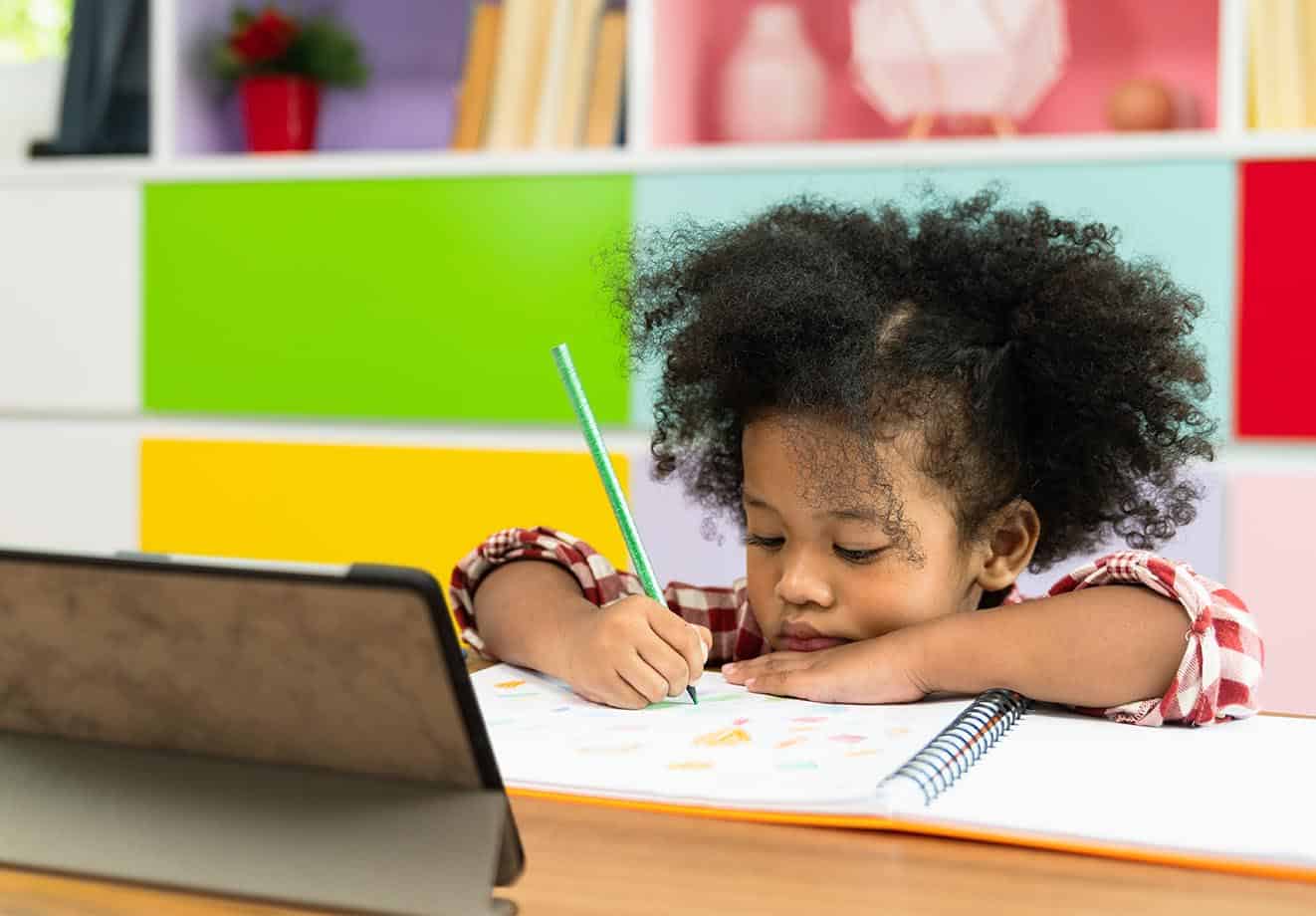 A child looks intently at and writes in a notebook with her chin on her desk