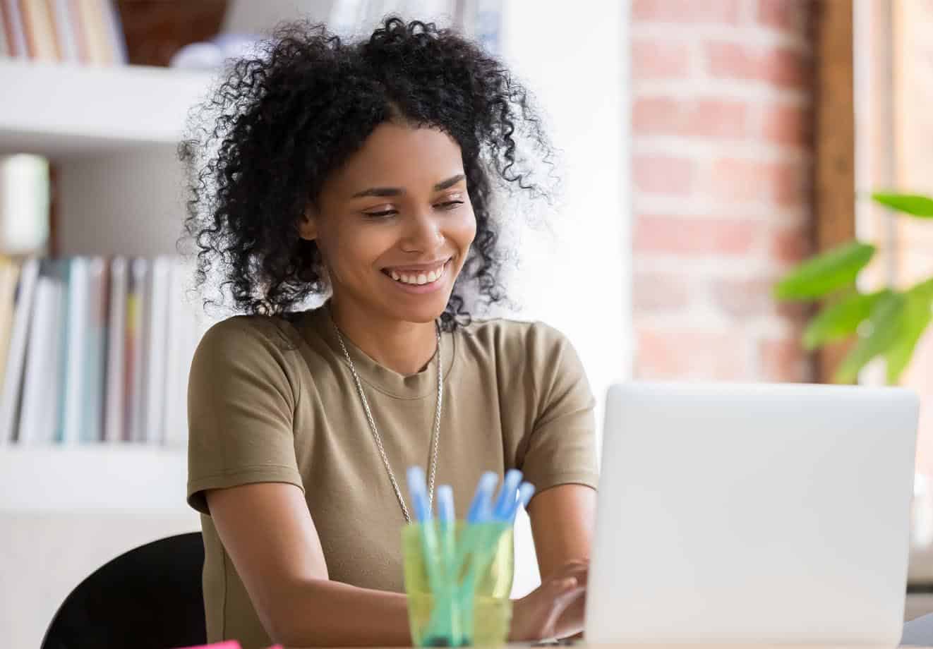 A teacher sits at a desk on a laptop with a bookshelf in the background