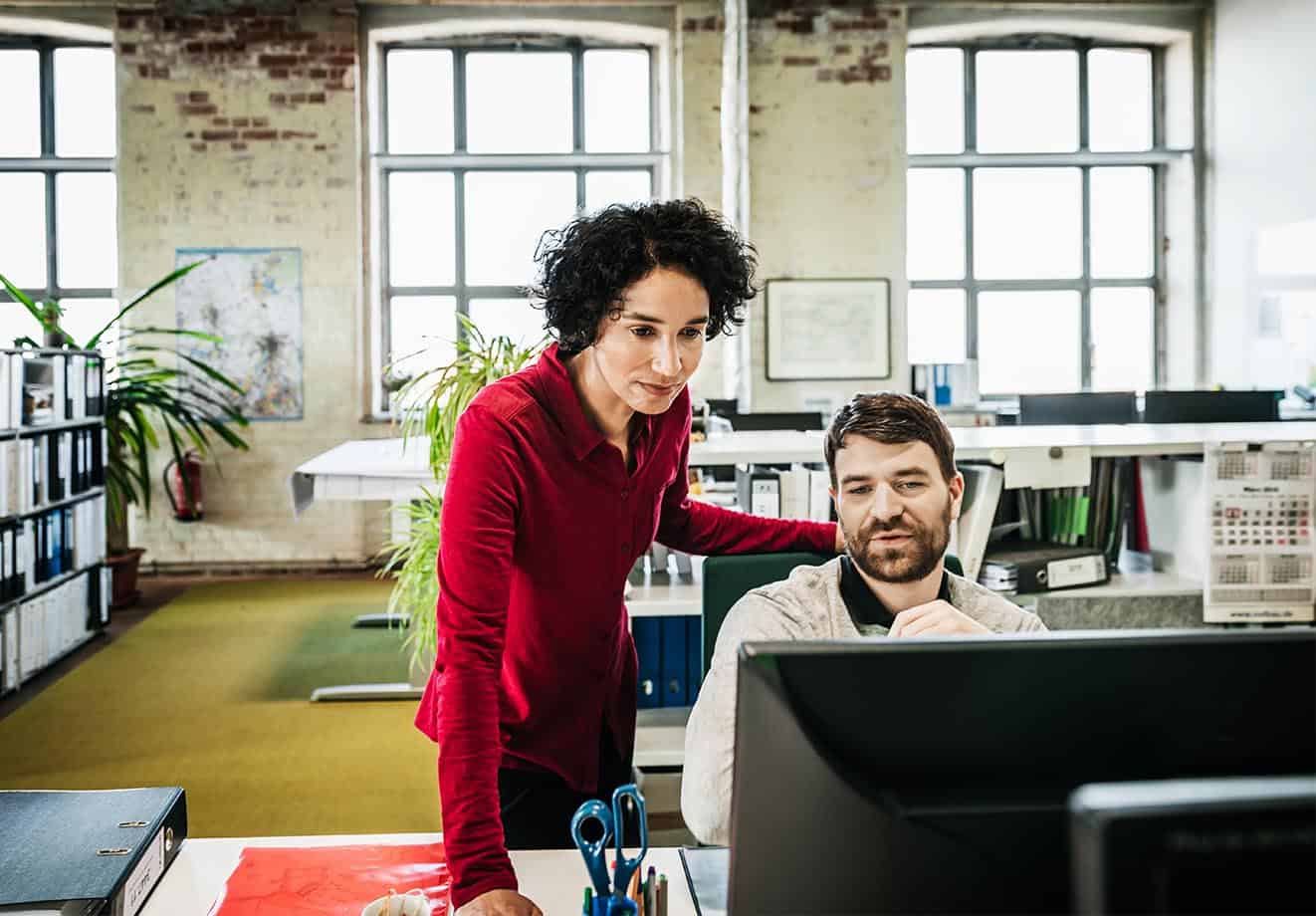 Man sitting at a desk looking at a computer while another co-worker looks over his shoulder