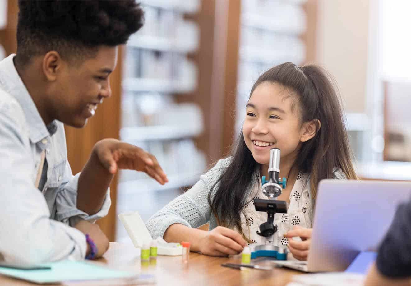 teacher and student smile at each other, while student uses microscope