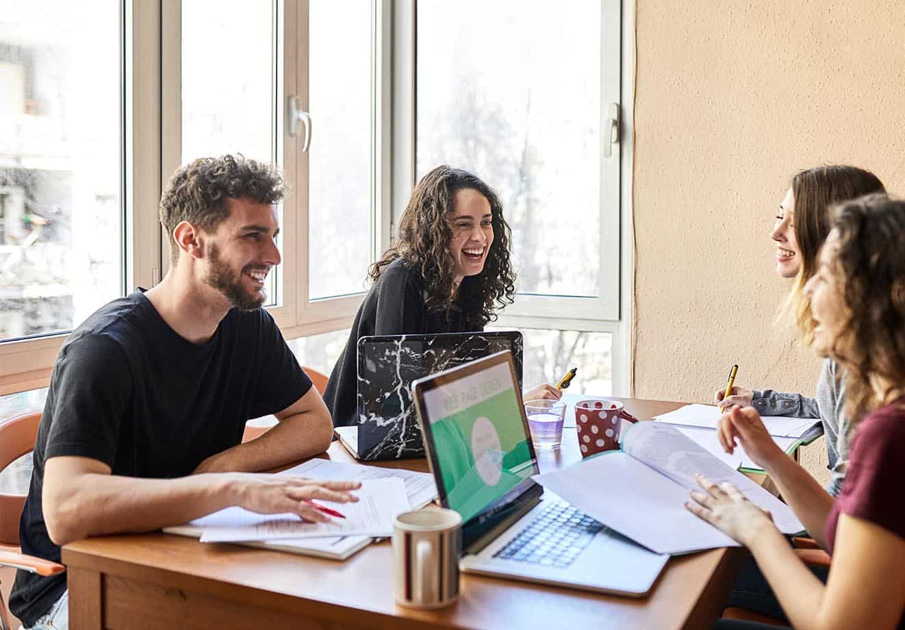 Students laughing while working at a table with laptops and books