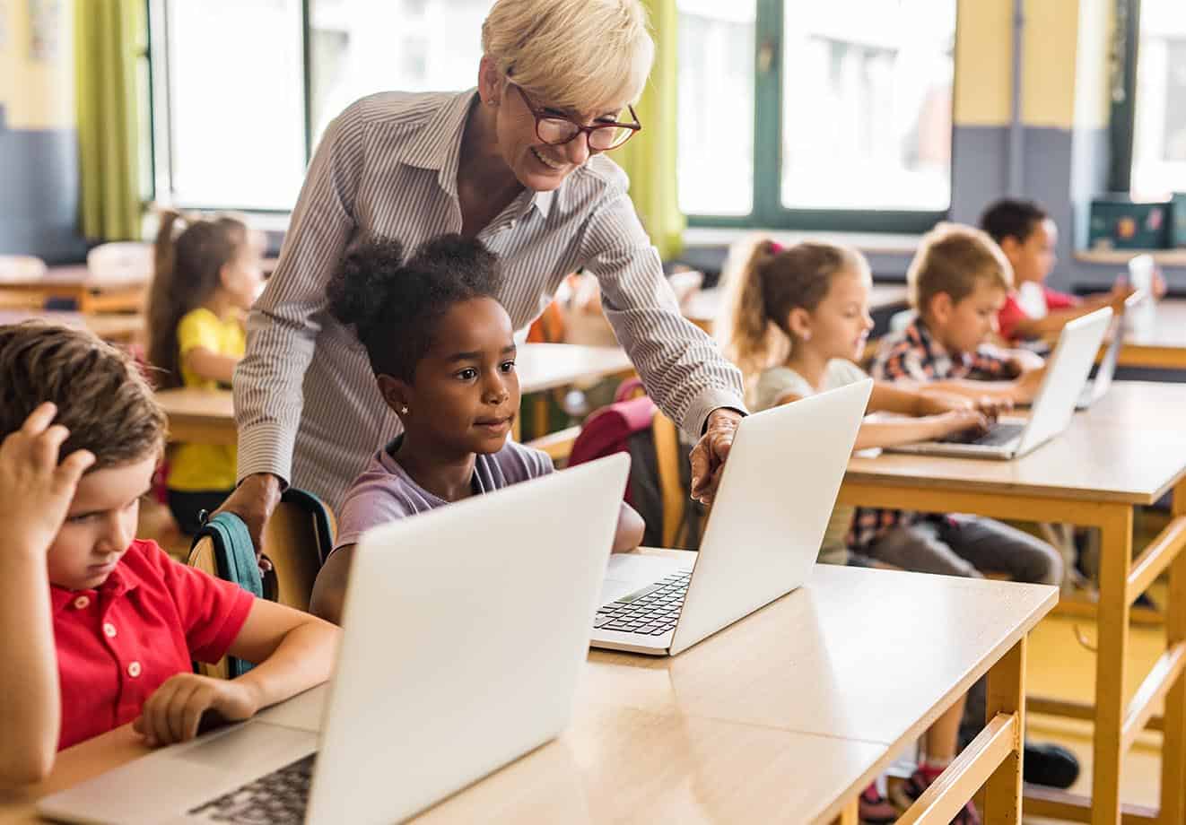 A teacher points to a student's laptop in a busy classroom.