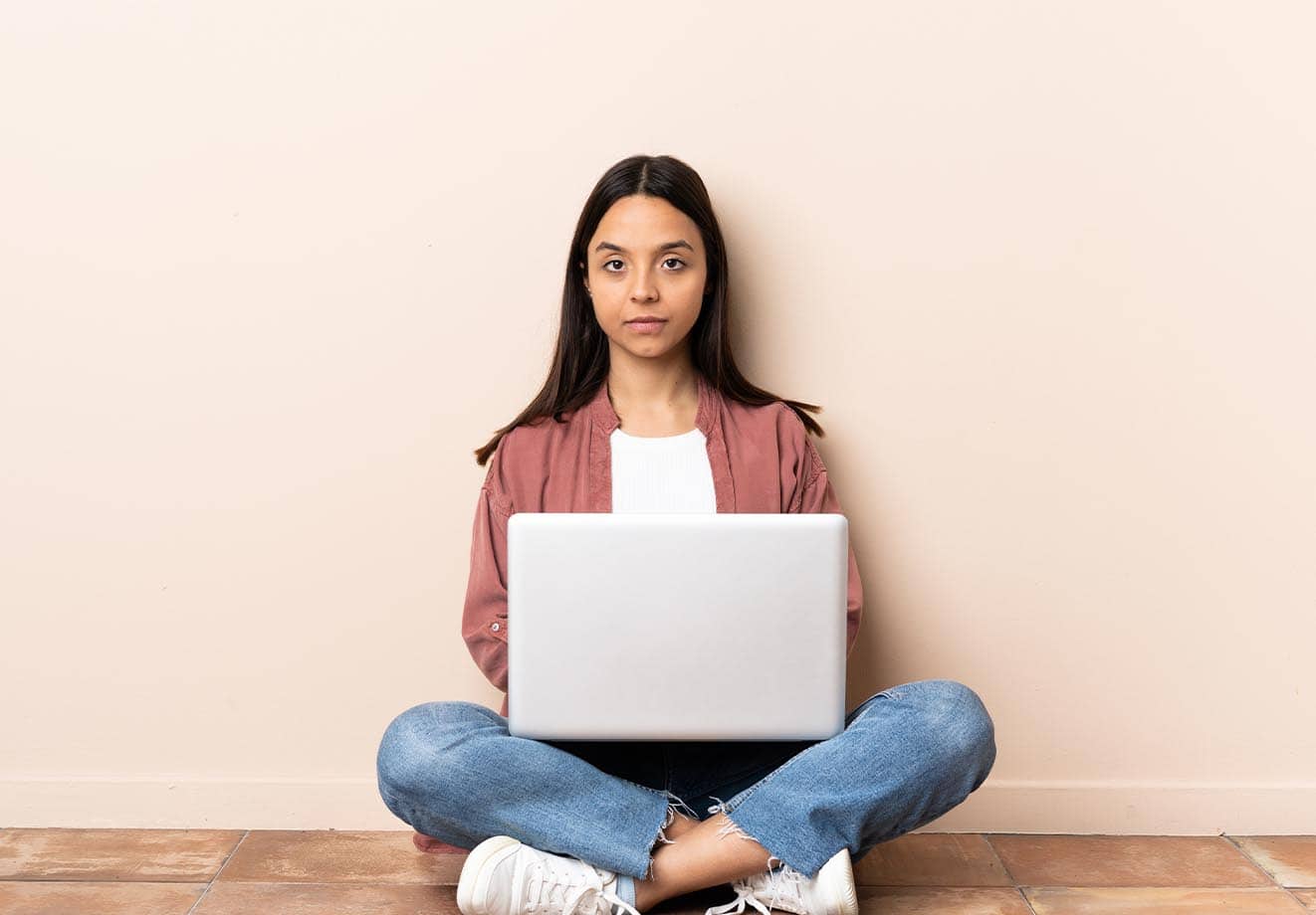 woman sitting cross-legged on the floor with a laptop on her lap looking toward the camera
