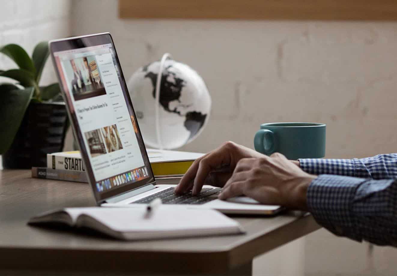 Close-up of hands on a laptop keyboard; background shows a glass globe, a coffee cup and a plant