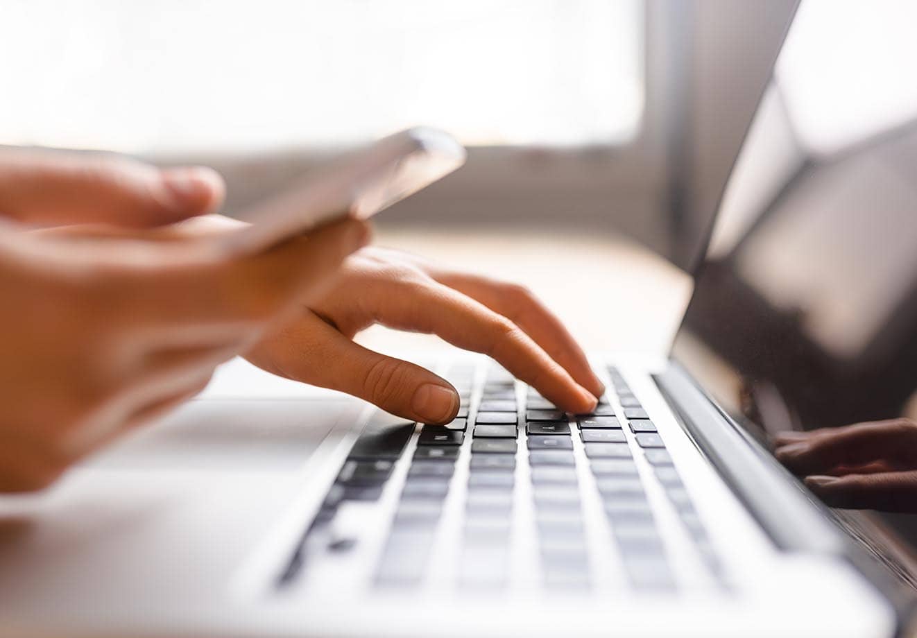 A person types on a keyboard with one hand while holding a cell phone in the other