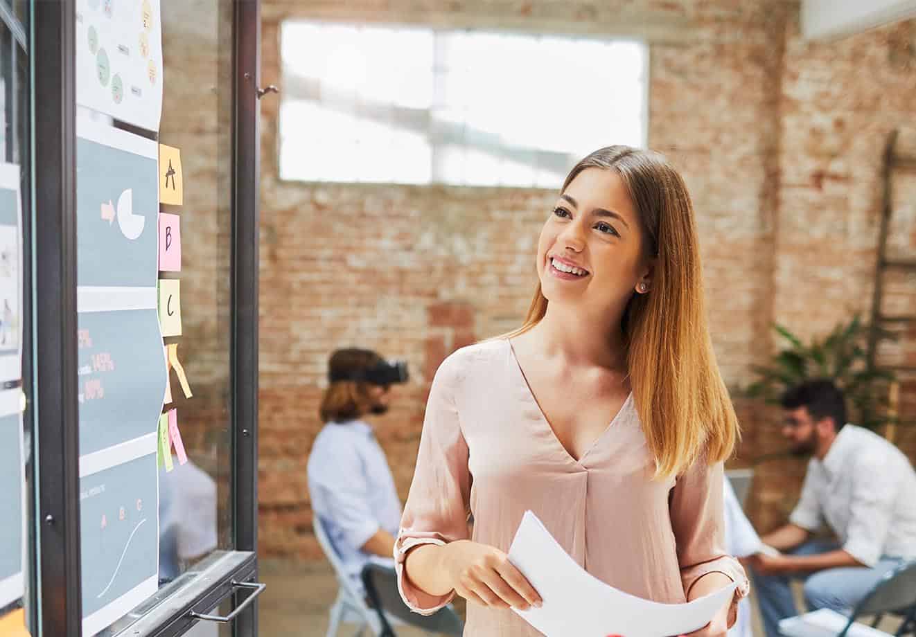 business woman looking at a whiteboard while holding a piece of paper in her hand
