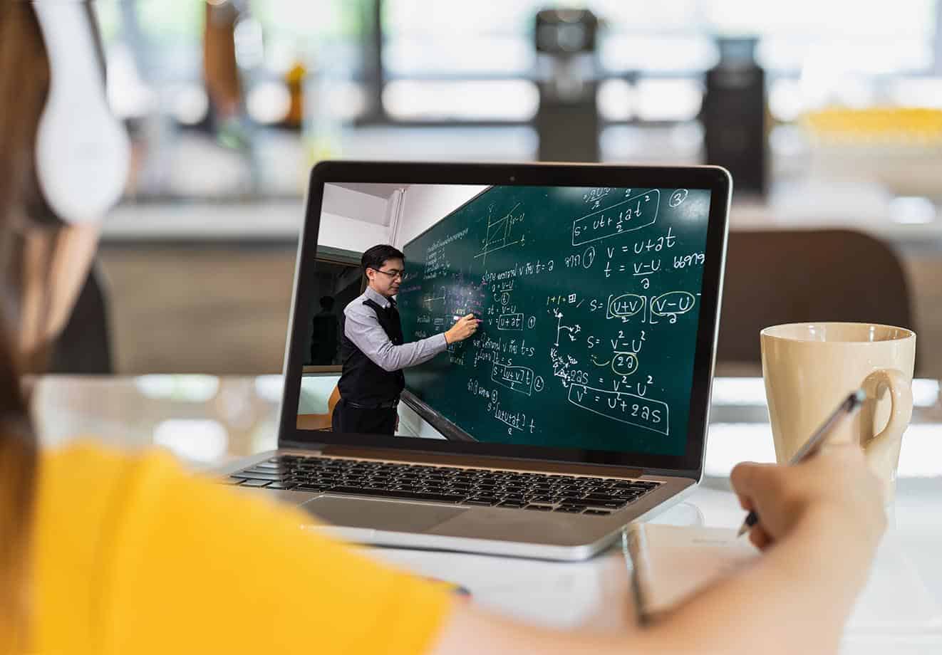 A student watches a math teacher on a laptop as he draws formulas on a chalkboard.