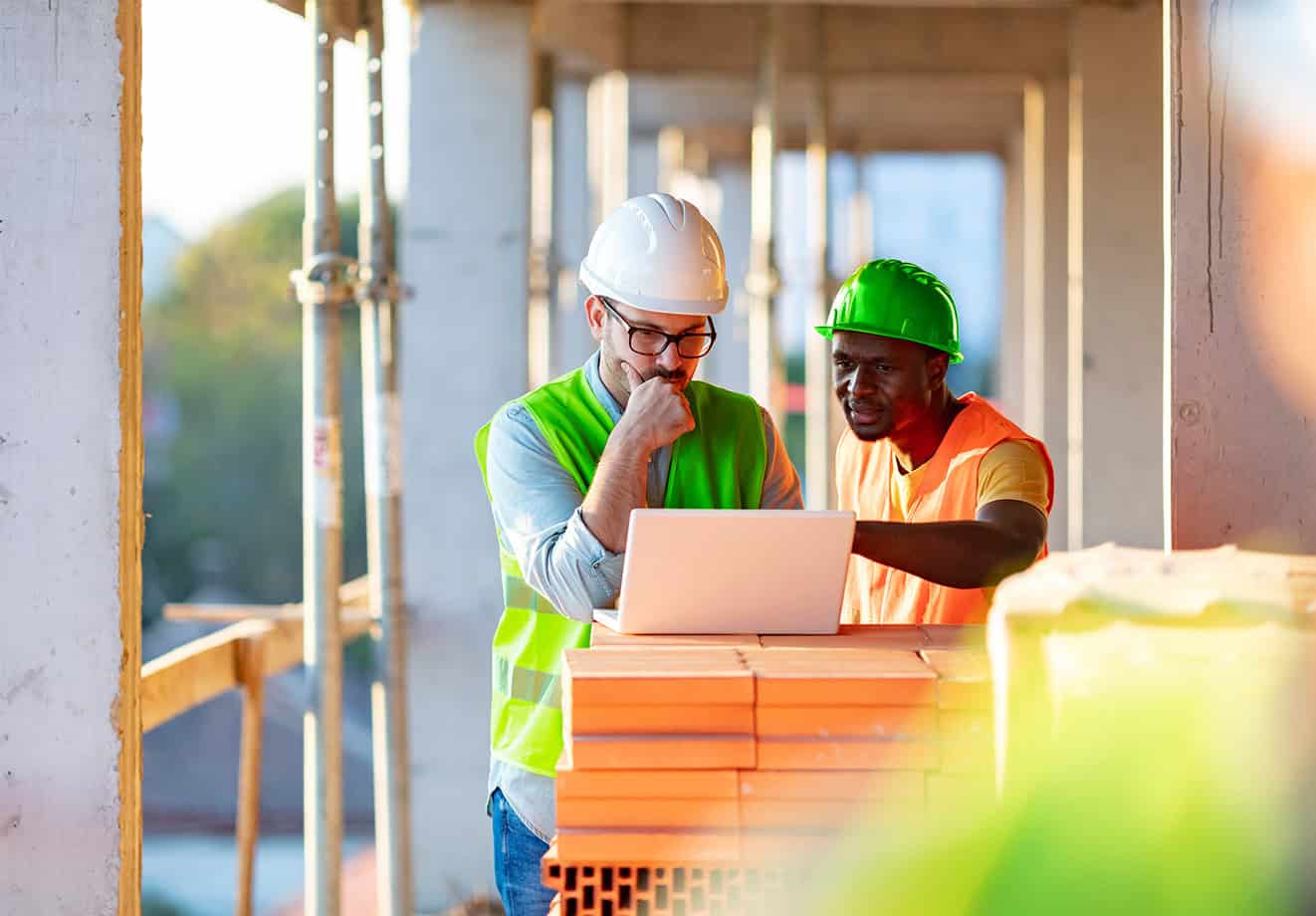 Two construction workers take training using a laptop while on a job site.