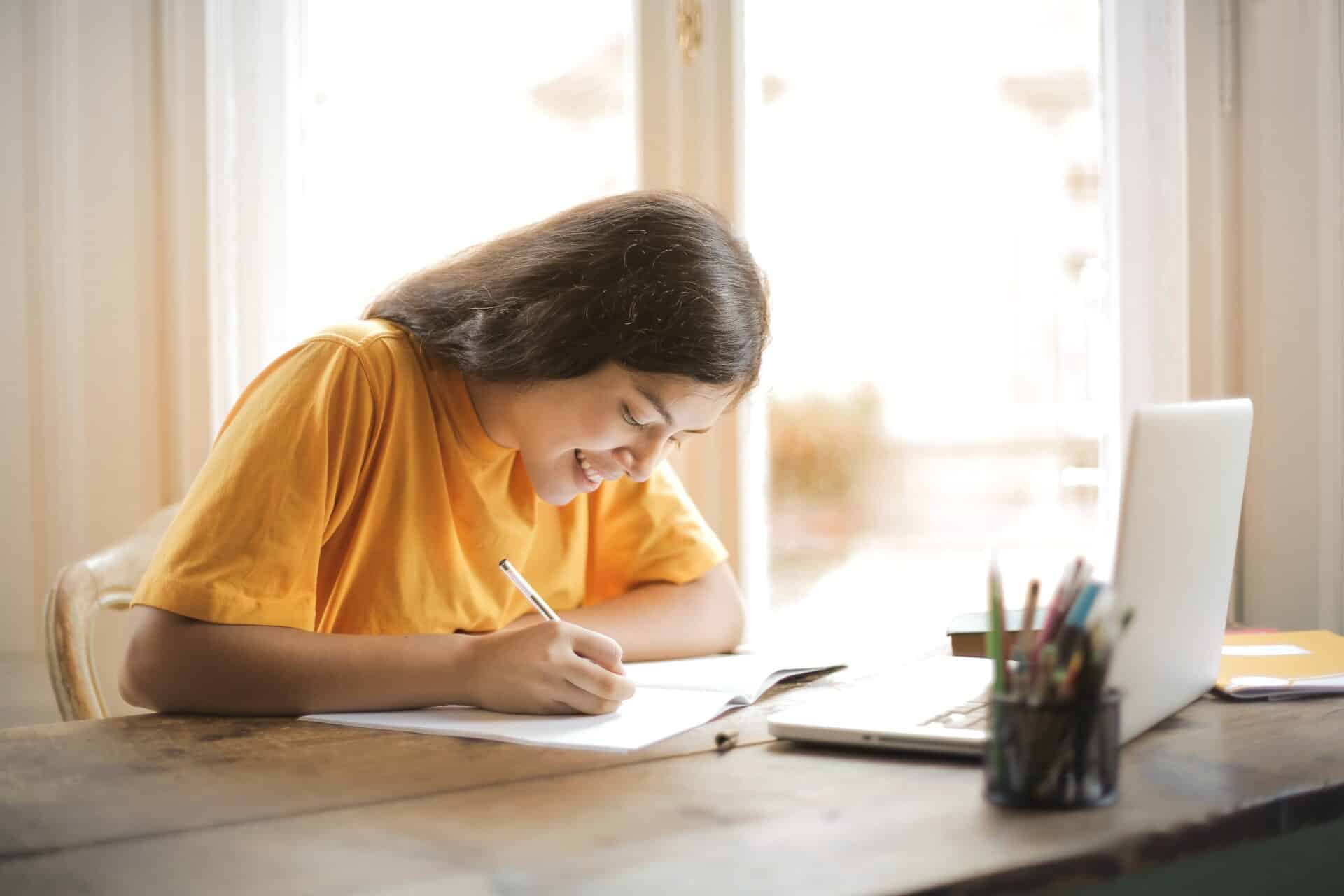 A student works in a notebook at a desk backlit by natural light from a window. There is a laptop on the desk.