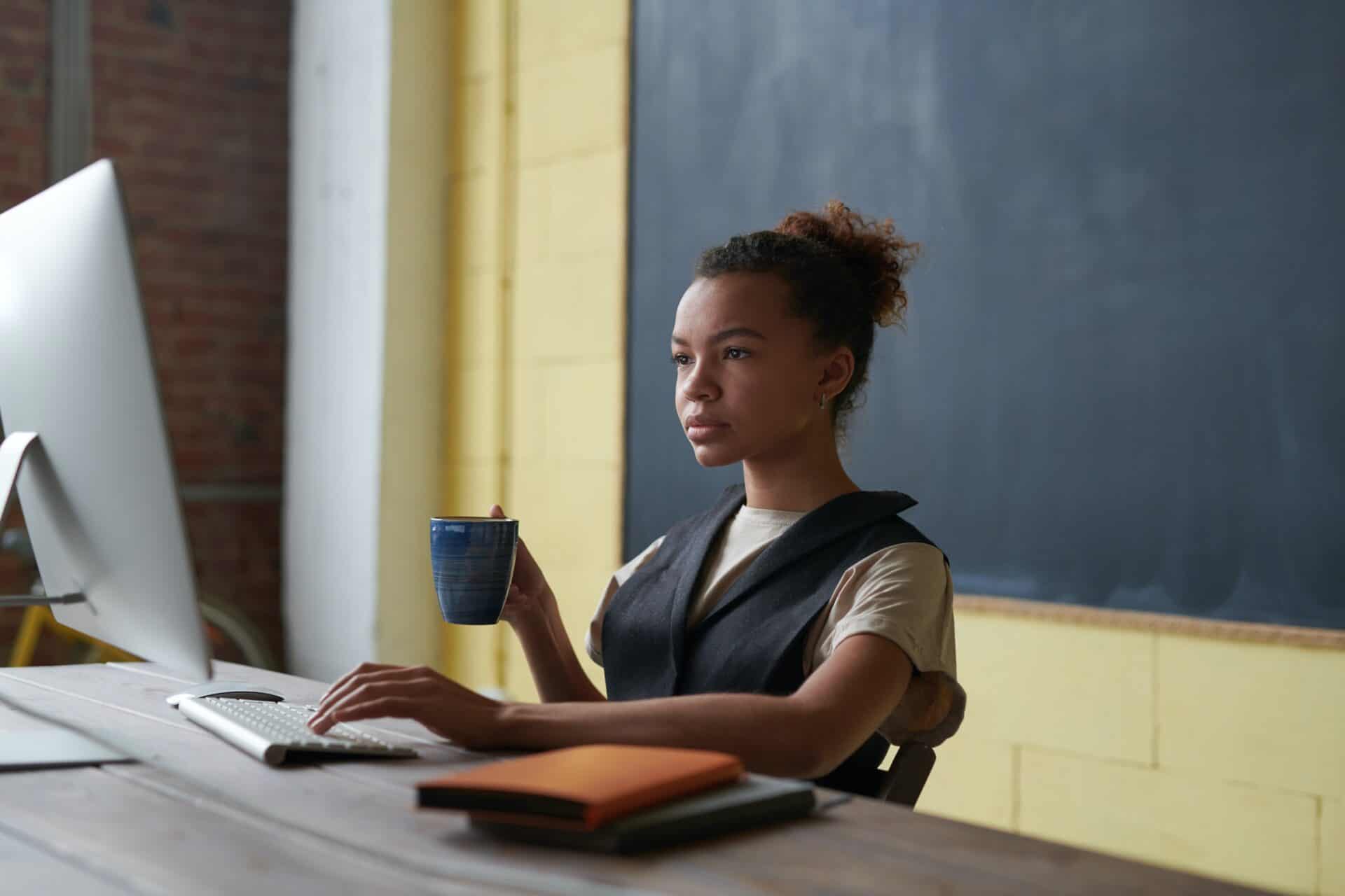 A teacher sitting at a computer with a coffee and a stack of notebooks