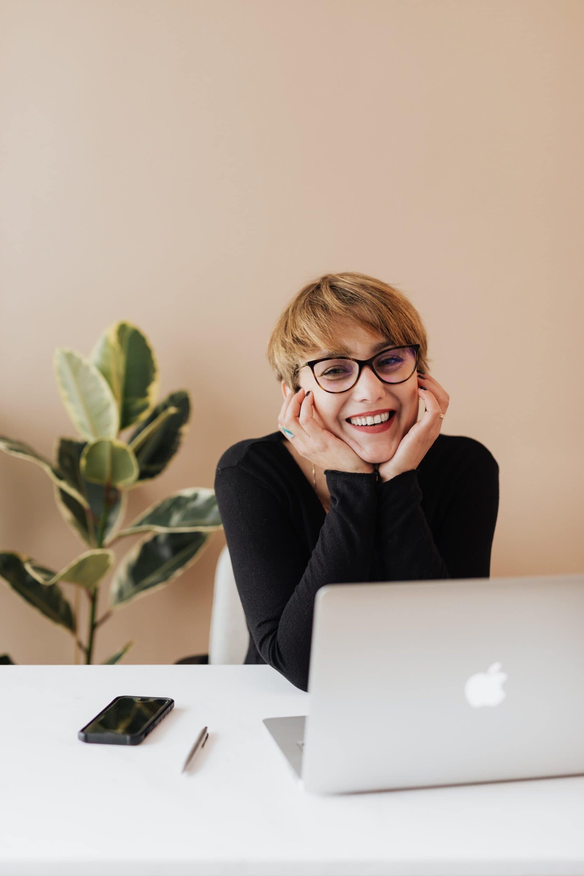 woman with short brown hair and black cat-eye glasses is smiling at the camera, with a laptop, cellphone and pen in front of her