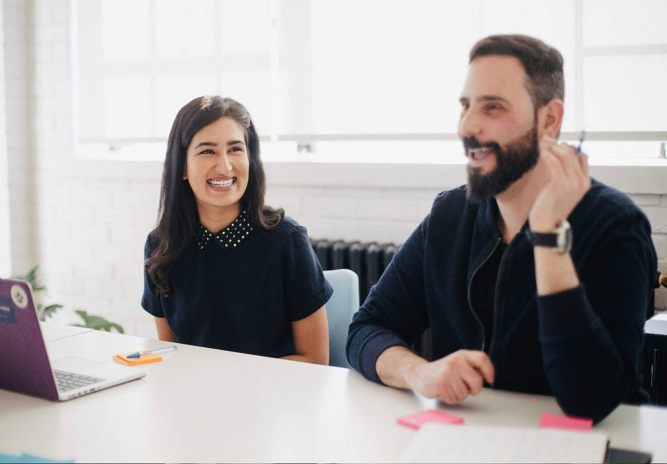 A man and a woman are sitting beside each other at a table. The man is talking to someone to someone on the other side of the table in front of them, and the woman is smiling.