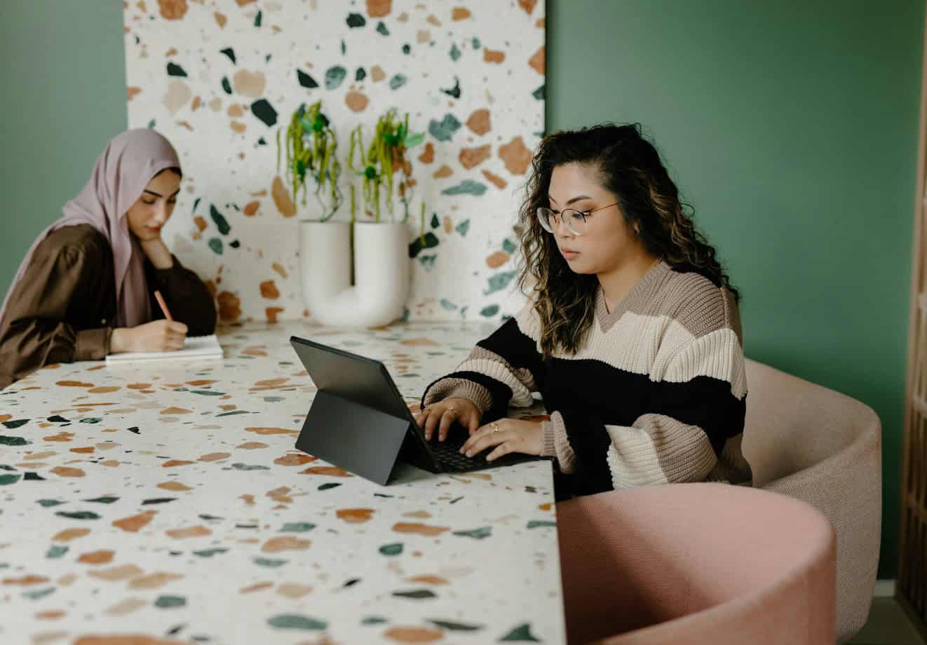 Two young women sitting at a long table working on their laptops