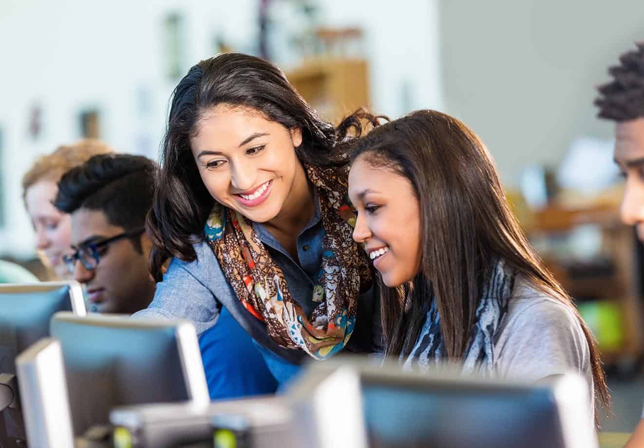 A student is looking at a desktop computer screen and a teacher is standing beside her to help her