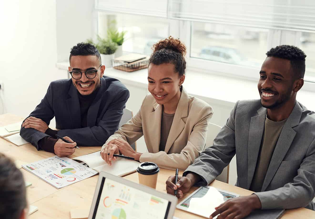 Three colleagues sitting around desk smiling and working on laptops