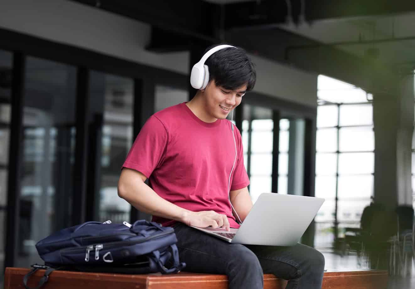 a boy sits on a bench and works on his laptop while wearing headphones
