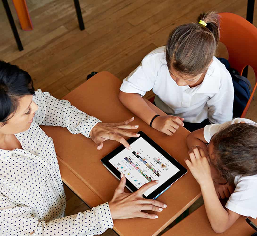 A teacher helping two young students use a tablet at their desk