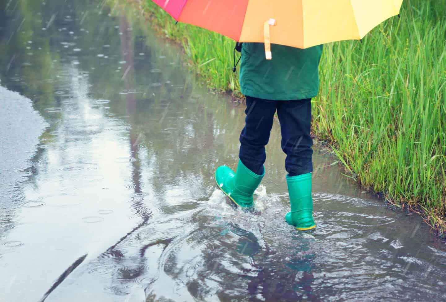Un enfant marche dans l'eau de pluie avec ses bottes en caoutchouc et avec son parapluie arc-en-ciel