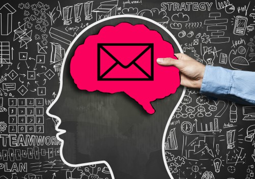 Hand holding paper cut out of a brain with an envelope icon. The brain cut out is poised over a chalk outline of a human profile, drawn on a blackboard, filled with complex mathematical figures.