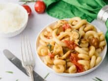 Mediterrane Halloumi-Pasta mit Zucchini und Tomaten serviert auf einem weißen Teller. Im Hintergrund ist ein Schüsselchen mit geriebenen Parmesan zu sehen. Davor liegt ein silbernes Besteck. Eine grüne Serviette ist bereit gelegt.