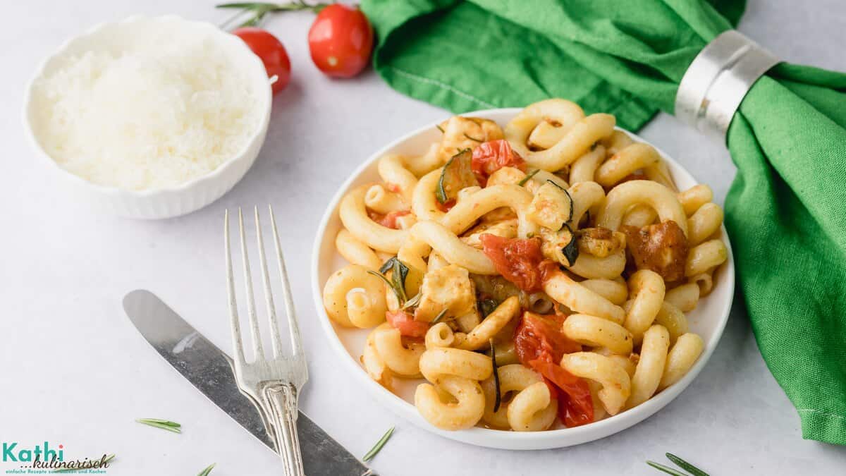 Mediterrane Halloumi-Pasta mit Zucchini und Tomaten serviert auf einem weißen Teller. Im Hintergrund ist ein Schüsselchen mit geriebenen Parmesan zu sehen. Davor liegt ein silbernes Besteck. Eine grüne Serviette ist bereit gelegt.