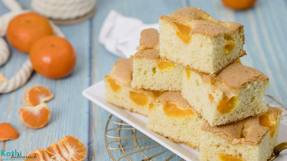 Dieser fruchtige Buttermilchkuchen mit Mandarinen ist in gleichmäßige Stücke geschnitten, die auf einem weißen Teller aufgestapelt sind. Im Hintergrund sind Früchte zu sehen.