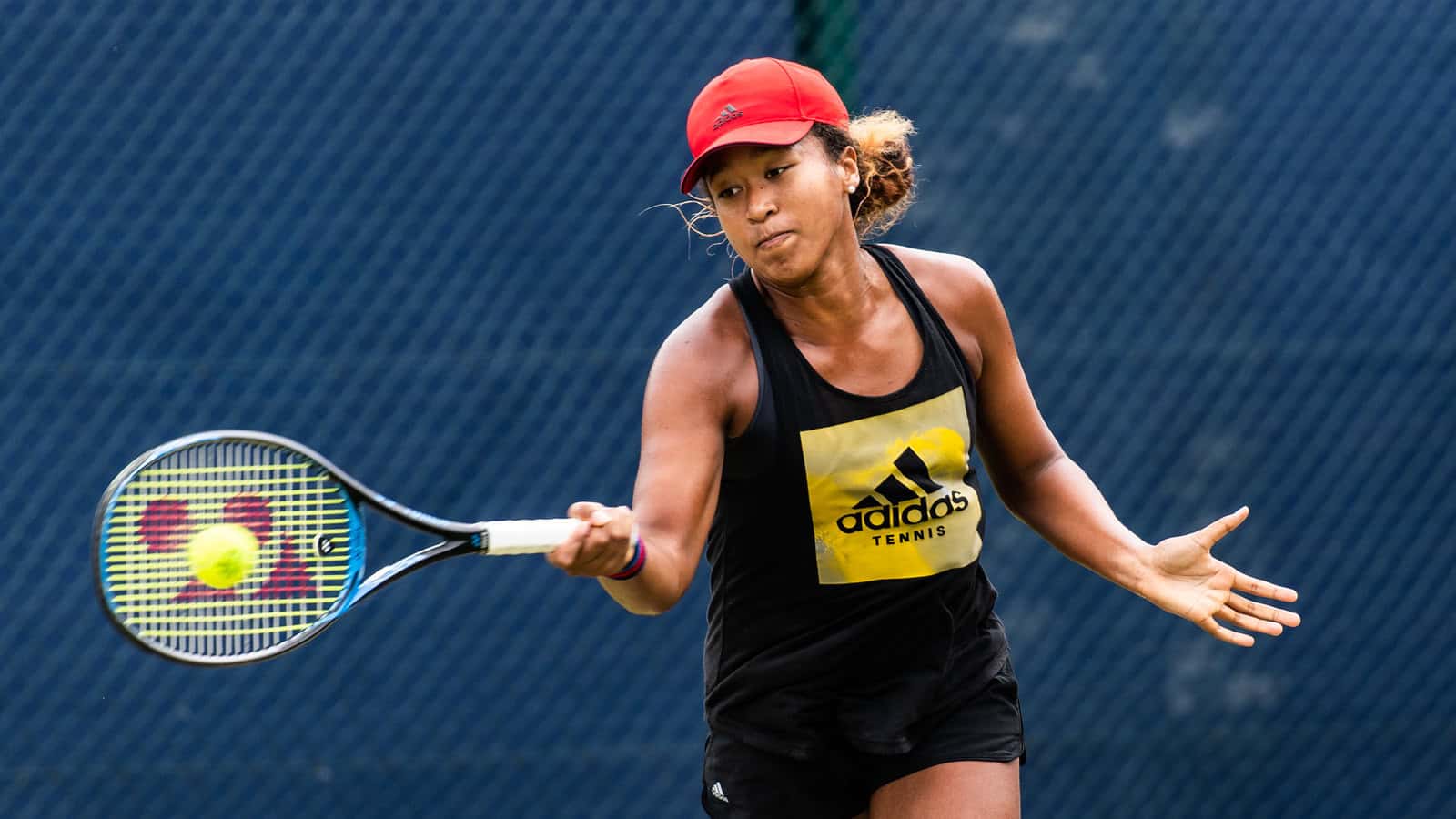 a Black woman wearing a black singlet with a yellow square and Adidas logo on the front, and a red cap. She is holding a tennis racquet and hitting a ball.