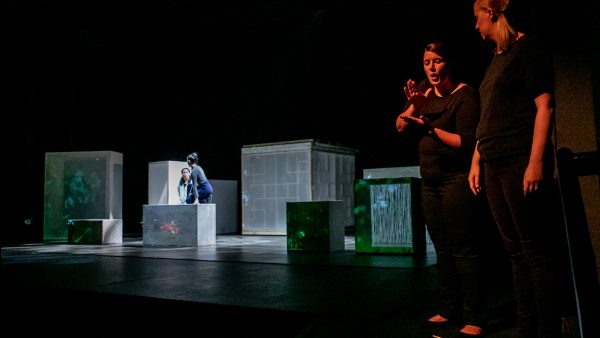 A dark stage with two Auslan interpreters in dim orange lighting, wearing black, in the foreground. In the background, two actors wearing casual clothes are brightly lit and surrounded by large metallic boxes.