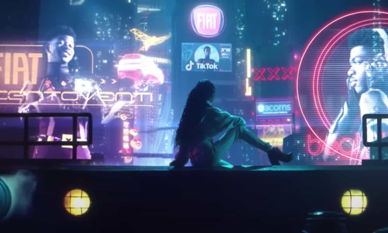 An image from Lil Nas X's 'Panini music video. A woman in the foreground is facing away from the camera, looking towards a futuristic cityscape full of large holographic advertisements featuring Lil Nas X