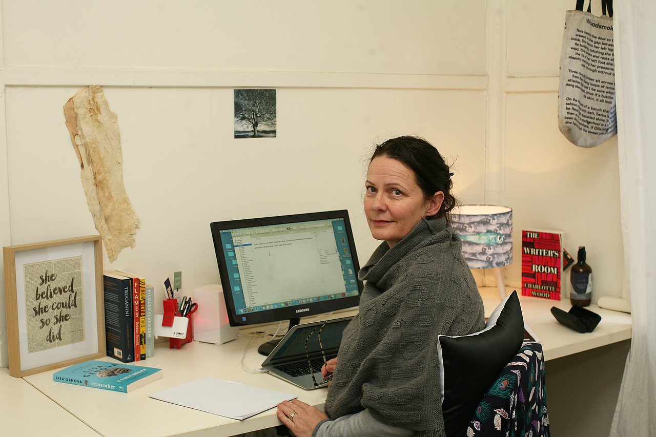Melissa Manning sitting at a white desk in a white room, turning to face the camera. Her hair is tied back, she is wearing a loose grey jumper and holding a pair of glasses. On the desk is a laptop connected to an external monitor, a lamp, books and a framed artwork with the words "she believed she could, so she did." On the wall is a large piece of bark and a small black and white photograph of a tree.