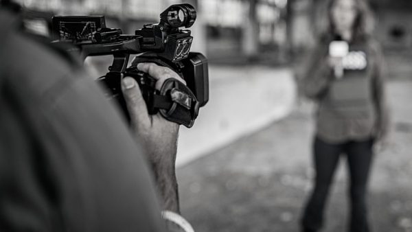 The shoulder and hands of a man holding a camera in the foreground, which is pointed at a white woman with long hair and a vest saying "press" standing in the background holding a microphone
