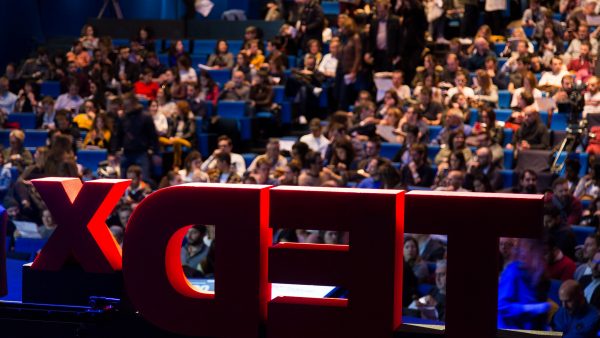 Large red 3D letters forming the TEDx logo, shot from behind on a stage facing toward an audience.