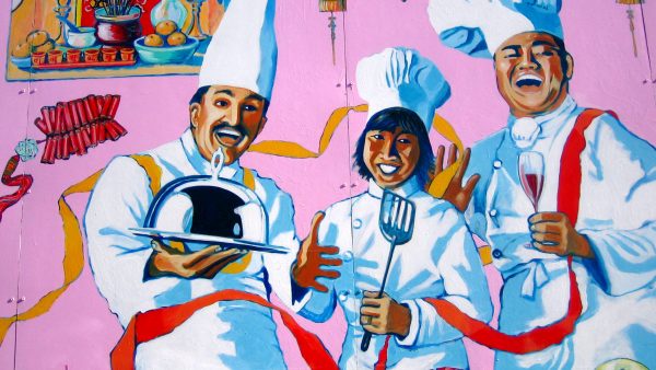 a painted mural of three smiling chefs - one is holding a cloche, one a spatula and another a glass of wine. Surrounding them on a pink background are various foods, lanterns and other items.