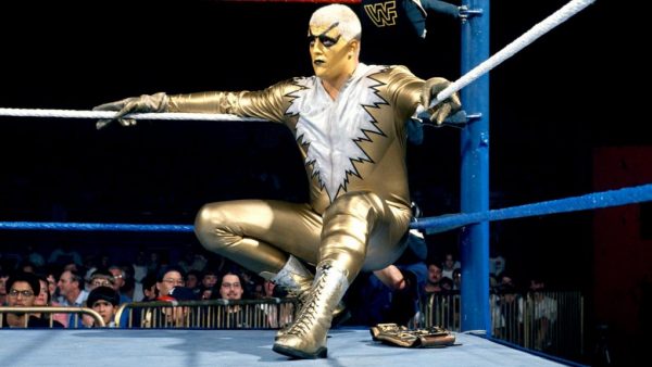 A wrestler wearing a gold body suit with a white mane-style pattern on the front, wearing gold face pain with black circles around the eyes, and short bleach-blond hair, leans against the ropes of a wrestling ring.