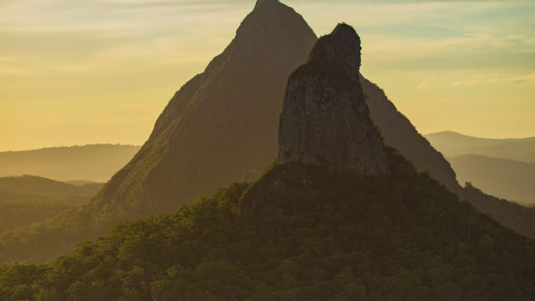 A long-range photograph of Mt Coonowrin, a large, thin jutting rock, with Mt Beerwah, a taller triangular mountain, behind it, in the early evening.