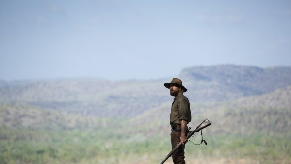 A man wearing a wide-brim hat and ragged clothing holds a rifle, against a backdrop of a semi-arid Australian outback landscape