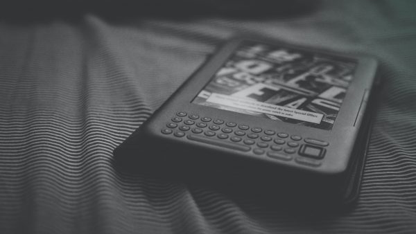 A soft-focus black and white photograph of an older-model Kindle lying on a bed