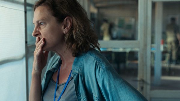 A still from the film 'Quo Vadis, Aida?'. Aida, a middle-aged woman with shoulder-length hair wearing a loose denim overshirt and a grey t-shirt, looks out a window with a concerned expression and her hand to her mouth.