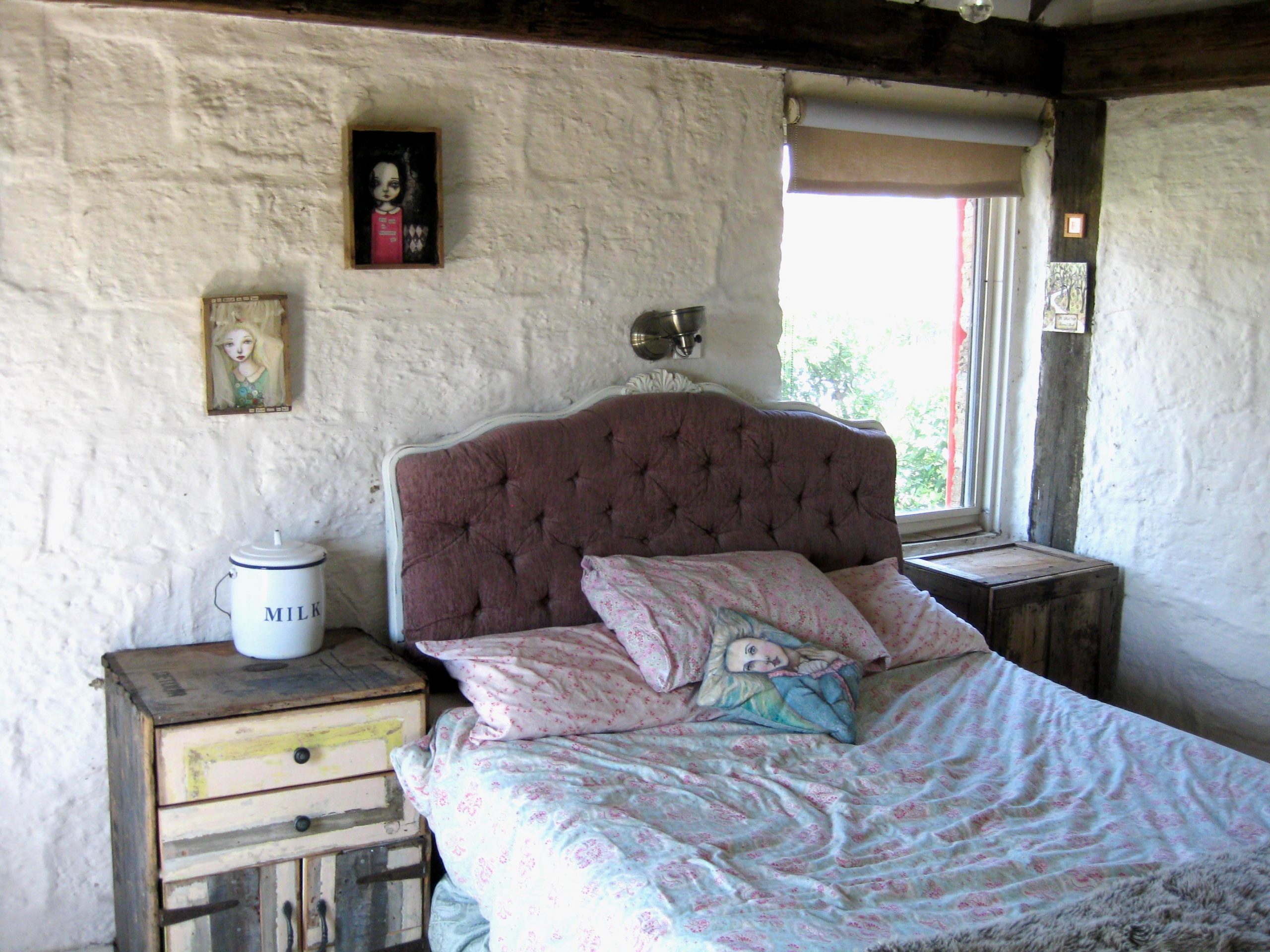 A double bed with blue and pink floral patterned bedsheets and pillows. On either side of the bed are timber crates repurposed into bedside tables, one of which has a large container labelled 'milk'. Paintings of Asphyxia's girl characters are on the wall and a throw pillow, and a large bush is visible out the window.