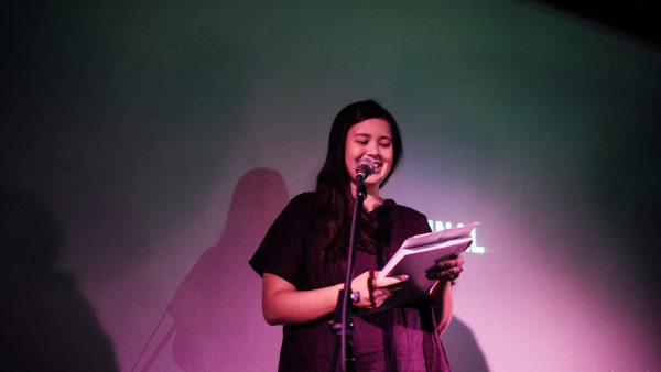 Leah Jing McIntosh wearing a black dress, standing behind a microphone with a pruple spotlight focused on her, holding a book in her hands.