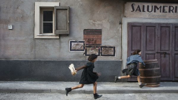 two young girls wearing dark coloured school dresses running along a dilapidated street in an Italian village.
