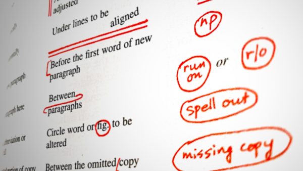 A close-up photograph of a book page, with proofreading marks in red handwritten text beside black typeset text outlining what each symbol means