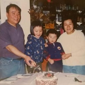 An old photograph of a middle-aged Chinese-Australian man and woman standing in a restaurant, with their two young children between them. The father and daughter are holding a knife above a birthday cake on the table in front of them.