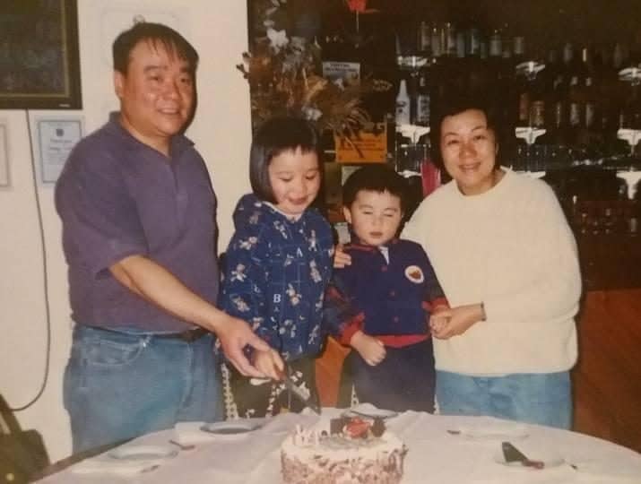 An old photograph of a middle-aged Chinese-Australian man and woman standing in a restaurant, with their two young children between them. The father and daughter are holding a knife above a birthday cake on the table in front of them.