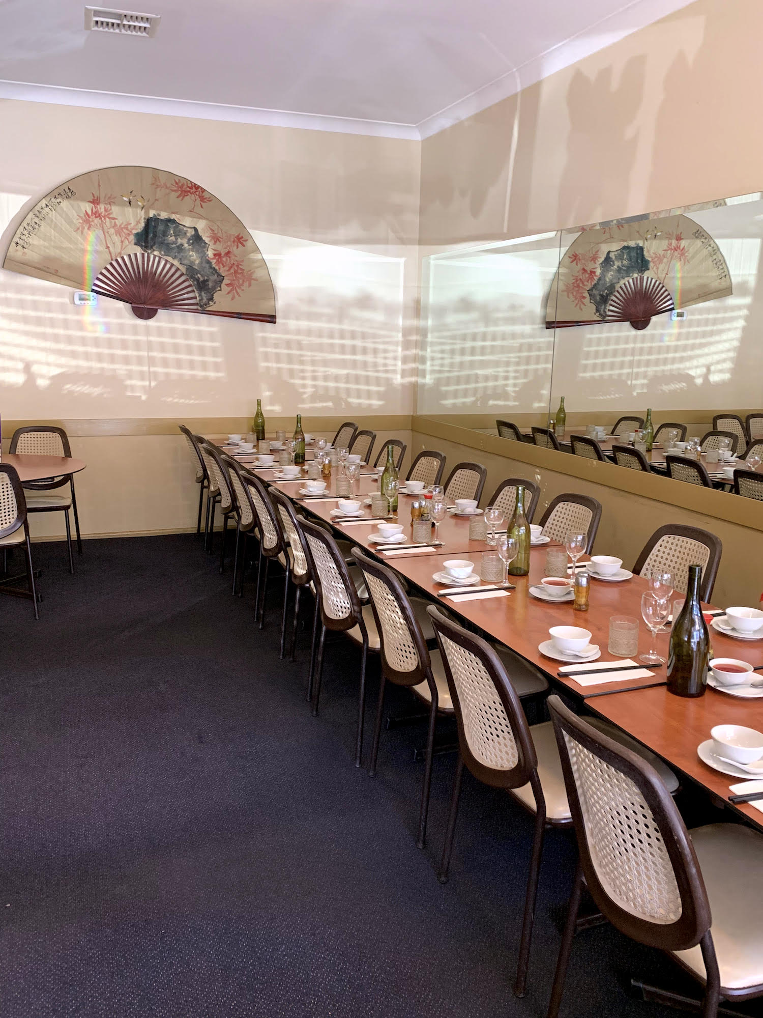 The interior of a Chinese restaurant. The floor has a dark blue carpet and a mirror runs along one wall next to a row of tables. On the far wall a large Chinese paper fan is mounted.