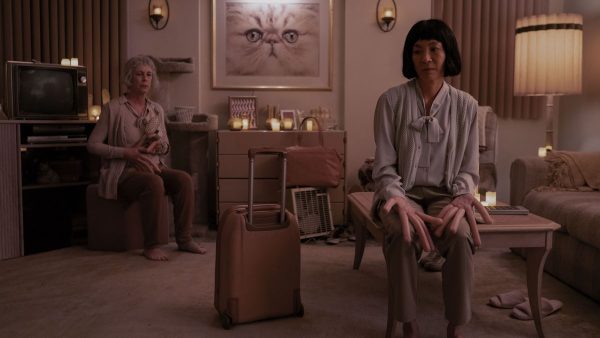 A scene from Everything Everywhere All At Once. Michelle Yeoh andJamie Lee Curtis' characters are sitting solemnly in a beige living room - their fingers are hot dogs.